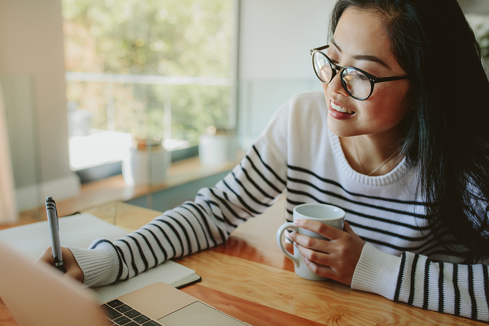 Woman writing notes in book holding cup of coffee in hand. Asian girl studying at home with laptop on table.
