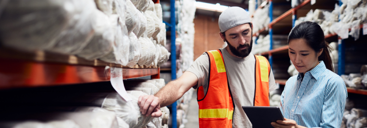 A man in a safety vest and a woman holding onto a digital tablet are both standing next to each other in a warehouse aisle.